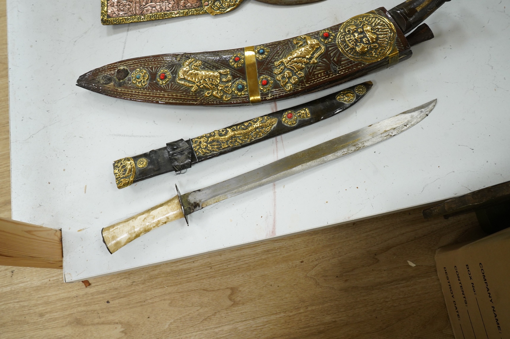 A decorative Indian axe, the large head inlaid with brass Hindu figures, together with a brass mounted North Indian kukri, another similar knife with bone handle, and a large shovel-shaped copper and brass item possibly
